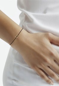 Juodas virvės apyrankė su auksiniu drugelio pakabučiu, lygia tekstūra, minimalistinis dizainas, dėvima ant rankos prieš baltą foną.