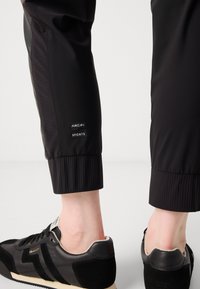 Black athletic pants with ribbed cuffs, featuring a Marc Cain tag. Paired with black and gray sneakers with a suede and leather mix.