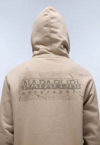 Sudadera beige con un logo gráfico texturizado en la parte posterior. Presenta una capucha con cordones y una textura de tela suave y lisa.