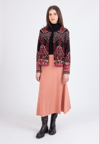 Vrouw in een zwarte turtleneck, een zwart-rode geblokte cardigan, een koraalroze midi-rok en zwarte kniehoge laarzen staat tegen een witte achtergrond.