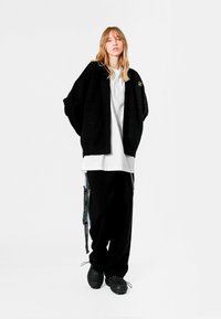 Cárdigan negro de punto oversize con un patrón texturizado, superpuesto a una camiseta blanca y pantalones negros de pierna ancha, combinados con calzado negro.