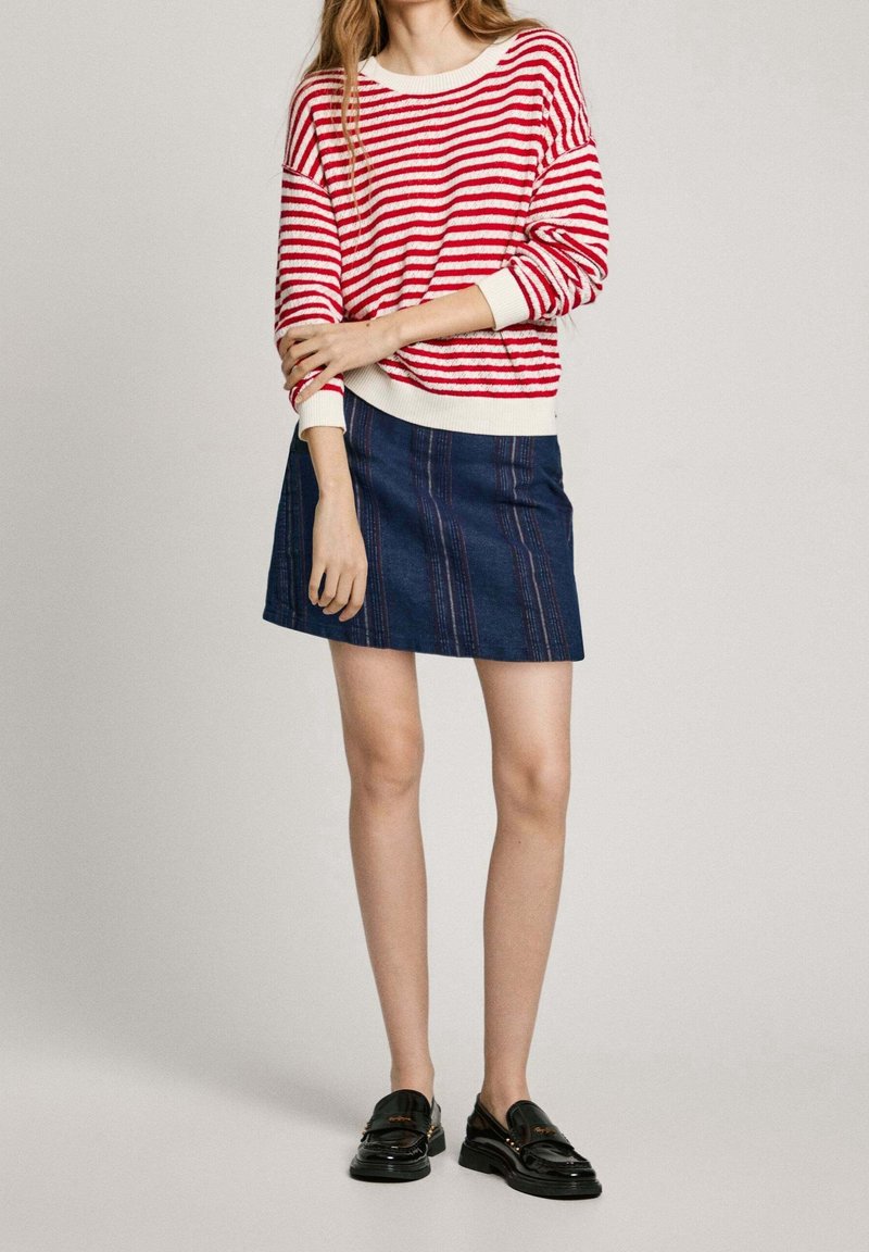 Femme portant un pull rayé rouge et blanc, une jupe bleu foncé et des mocassins noirs, debout les bras croisés devant un fond uni.