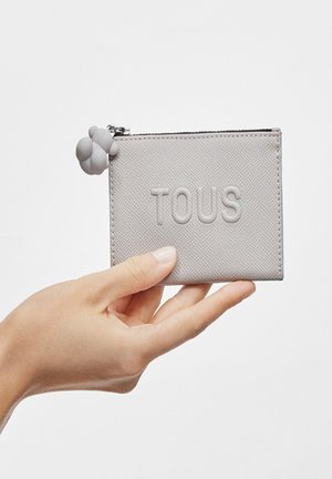 Cartera texturizada gris con el logo embossado "TOUS", cierre de cremallera y un pom-pom decorativo de silicona gris adjunto a la cremallera.