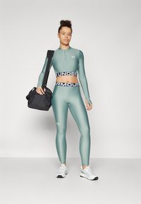 Strak figuurvormend sportoutfit in gedempt groen met een zwarte "Under Armour" tailleband, gecombineerd met een zwarte tas en witte sneakers.