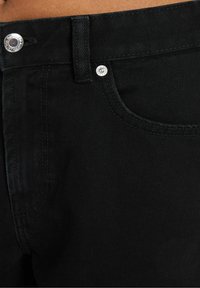 Černé džínové kalhoty z denimu s hladkou texturou, přiléhavým střihem a stříbrnými kovovými knoflíky na pasu a kapsách.