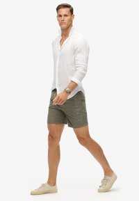 Camicia bianca in lino con maniche arrotolate, pantaloni corti verde oliva con il testo "SUPERDRY" sul lato, sneakers beige chiaro e un orologio argento.