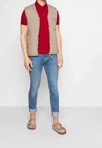 Gilet matelassé beige sur un polo rouge, associé à un jean bleu clair à revers et des sandales grises à boucles. Tissu lisse et coupe décontractée.
