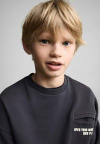 Tmavo siva mikica s krojem okoli vratu, z sprednjim žepom in belim napisom "ODPRETE SVOJ UM NOV PLL." Kratke, razmršene svetlo rjave lase in modre oči.