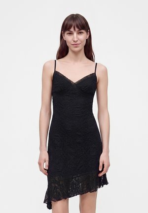Jeune femme aux cheveux bruns et raides portant une robe noire en dentelle sans manches, avec de fines bretelles et un ourlet asymétrique.