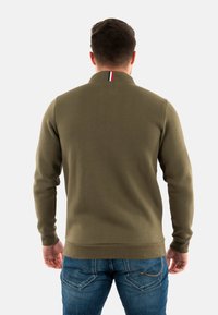Felpa verde oliva con colletto alto, polsini e orlo a coste. Presenta un dettaglio a righe sul colletto posteriore. Indossata con jeans blu.