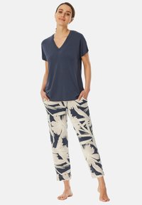 Marineblaue V-Ausschnitt-Bluse mit kurzen Ärmeln, kombiniert mit cremefarbenen Hosen, die ein großes, marineblaues und weißes tropisches Blättermuster aufweisen, gefertigt aus weichem, leichtem Material.