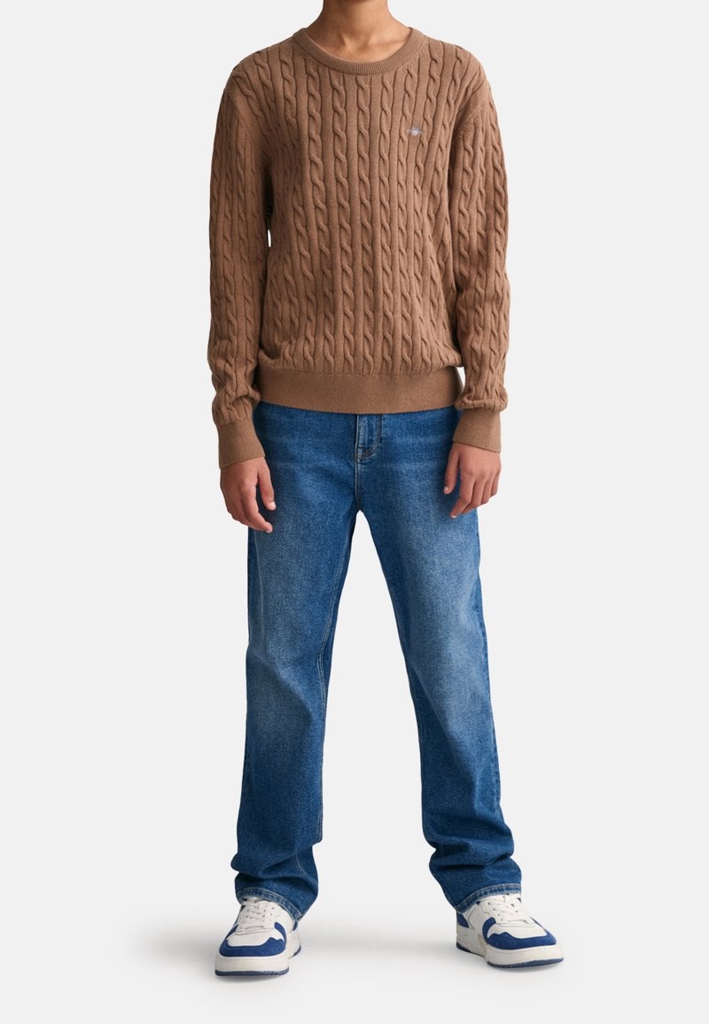 Suéter marrón de punto con cuello redondo, combinado con jeans azul claro. Zapatillas blancas con detalles azules completan el conjunto.