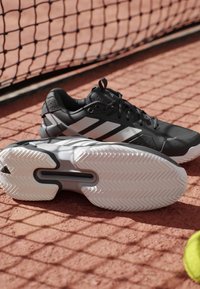Czarne buty sportowe z szarymi akcentami, wyposażone w teksturowaną gumową podeszwę i charakterystyczny wzór w paski. Idealne do sportów na kortach.