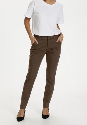 Kobieta stoi z rękoma w kieszeniach, ubrana w białą koszulkę z krótkim rękawem, brązowe dopasowane spodnie i czarne szpiczaste szpilki.