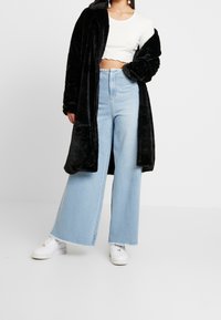 Mustat tekoturkistakkia, valkoinen lyhyt toppi, vaaleansiniset leveälahkeiset farkut ja valkoiset tennarit. Farkuissa huomattava risatorvi.