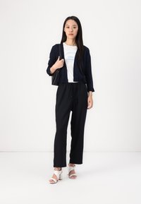 Pantaloni neri con elastico in vita, abbinati a una t-shirt bianca con testo blu e un cardigan blu navy. Sandali con tacco bianchi. Borsa nera.