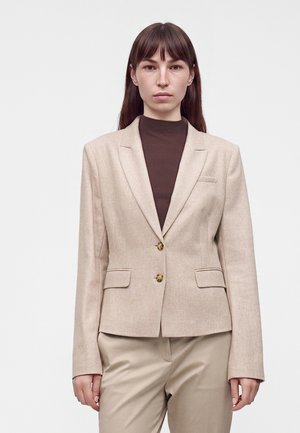 Junge Frau mit langen braunen Haaren, die einen beigen Blazer über einem dunkelbraunen Rollkragenpullover und beigen Hosen trägt, steht vor einem einfarbigen hellen Hintergrund.