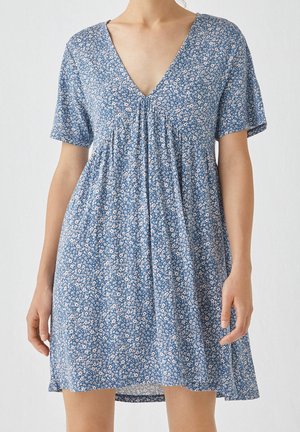 Robe bleue à manches courtes avec un petit motif floral blanc et orange, col en V, coupe ample portée par une personne debout.
