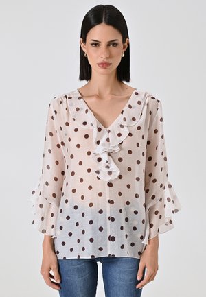 Camicia leggera e trasparente con pois marroni, scollatura a V, fronte arricciato e maniche a campana. Il tessuto sembra morbido con una texture leggermente trasparente.