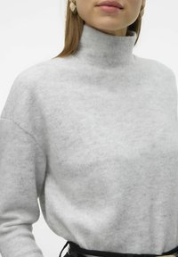Pull en col roulé gris clair avec une texture douce, coupe ample et épaules tombantes. Accentué d'une ceinture noire.