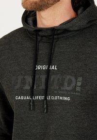 Mörkgrå hoodie med en framsida designad med ordet "UNITD" och mindre text. Materialet verkar mjukt med en lätt texturerad yta.