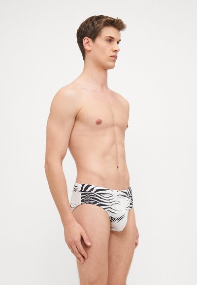Slipuri de baie pentru bărbați, de culoare albă, cu un model în dungi de zebră negre. Prezintă un tiv cu branding subtil și un design ajustat.