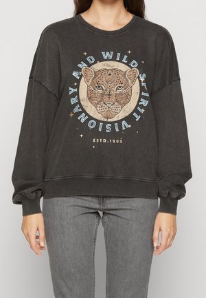 Osoba w ciemnoszarej, oversize'owej bluzie z detaliczną twarzą lamparta i napisem "VISIONARY AND WILD SPIRIT" wokół niej, w połączeniu z szarymi dżinsami.