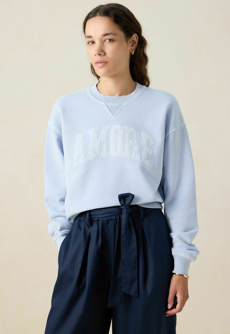 Lichtblauwe sweatshirt met "AMORE" in verhoogde letters, gecombineerd met donkerblauwe broeken met een gestrikt taille. Zachte stof, ontspannen pasvorm.