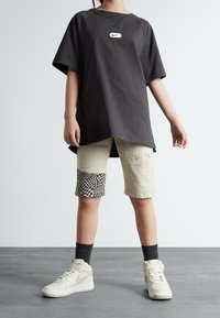 Camiseta negra de gran tamaño con un logo blanco de Nike, pantalones cortos de patchwork beige con un patrón de tablero de ajedrez y zapatillas blancas con acentos verdes.