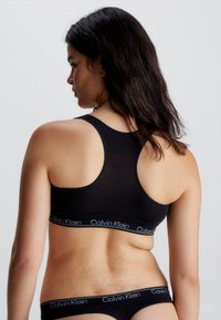 Svart bomull sports-bh med racerback-design og en myk elastisk kant som har "Calvin Klein"-logoen i hvitt. Glatt tekstur.