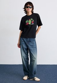 Černé tričko s barevným ovocným motivem, volnější střih, krátké rukávy. V kombinaci se světlými modrými džíny a bílými teniskami s černými akcenty.