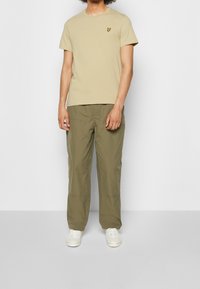 Beżowa koszulka z okrągłym dekoltem z małym żółtym logo ptaka, zestawiona z jasnymi oliwkowozielonymi spodniami typu cargo. Całość dopełniają białe trampki.