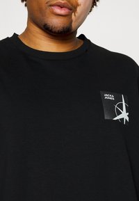 T-shirt nera in cotone con un piccolo logo patch bianco e nero sul petto sinistro con un design geometrico. Stile classico a girocollo.