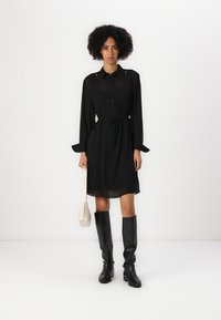 Svart, gjennomsiktig knappedress med lange ermer, innsnørt midje og sidelommer. Kombinert med knelange svarte støvler og en liten hvit veske.