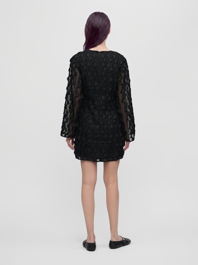 Modèle vu de dos, portant une mini-robe noire texturée à manches longues et des chaussures plates, sur un fond blanc uni.