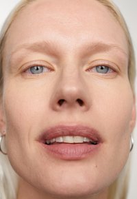 Gros plan sur un visage à la peau claire, avec des yeux bleus, un léger blush sur les lèvres et une texture lisse. Les cheveux sont blonds et raides, agrémentés de boucles d'oreilles en argent.