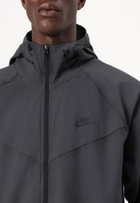 Nike hooded jack in donkergrijs materiaal, met een volledige rits, verstelbare koorden en een subtiel logo op de linkerborst. Soepele textuur met naaddetails.