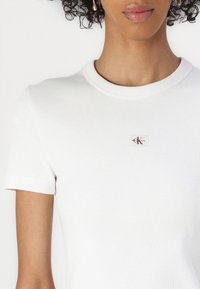 T-shirt bianca a coste con maniche corte, scollatura rotonda e un piccolo patch quadrato con testo rosso e nero sul petto.