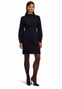 Schwarzes Strickkleid mit langen Ärmeln, Rundhalsausschnitt und taillierter Silhouette. Glatte Textur, getragen mit glänzenden schwarzen High Heels und transparenten Strumpfhosen.