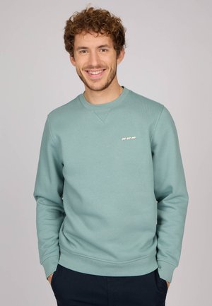 Sweatshirt vert menthe en tissu doux, avec un col rond, des manches longues et un petit motif blanc sur la poitrine.