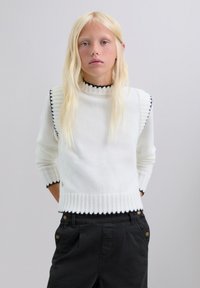 Suéter blanco de punto con dobladillo y cuello acanalados, que presenta acentos en negro a lo largo de los hombros y puños. Se lleva con pantalones cargo negros.
