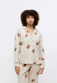 Lys beige pysjamasett med bamseprint, laget av myk stoff. Knappeløs skjorte med lange ermer og matchende bukser.