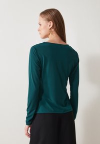 Hosszú ujjú felső mély teal színű anyagból, sima textúrával és kerek nyakkal, laza szabással és minimalista dizájnnal.