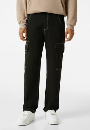 Pantalon cargo noir avec coutures blanches porté avec des baskets blanches et un sweat beige, montré de la taille aux pieds.