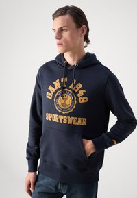 Navy blue hoodie made of cotton, featuring a kangaroo pocket, drawstring hood, and gold printed branding and logo on the front.