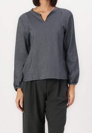 Vrouw draagt een blouse met lange mouwen in marineblauw en wit met verticale strepen en een V-hals, gecombineerd met losse donkergrijze broek.