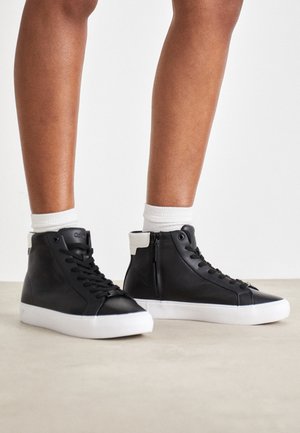 Persona con zapatillas negras de caña alta con suelas blancas y calcetines blancos hasta el tobillo, de pie sobre un suelo de color claro contra un fondo liso.