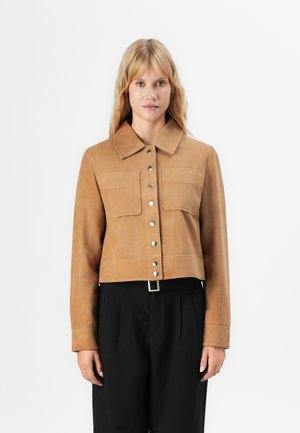 Woman with shoulder-length blonde hair wearing a tan buttoned jacket with flap pockets and black high-waisted trousers.