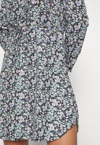 Robe noire avec un imprimé floral dense de petites fleurs blanches, violettes et jaunes, taille cintrée avec un détail à nouer, manches longues et une fente latérale dévoilant la cuisse.