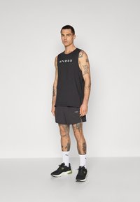 Zwarte mouwloze top met "HYROX" opdruk, donkere shorts, zwarte hardloopschoenen met groene accenten en witte crew sokken.